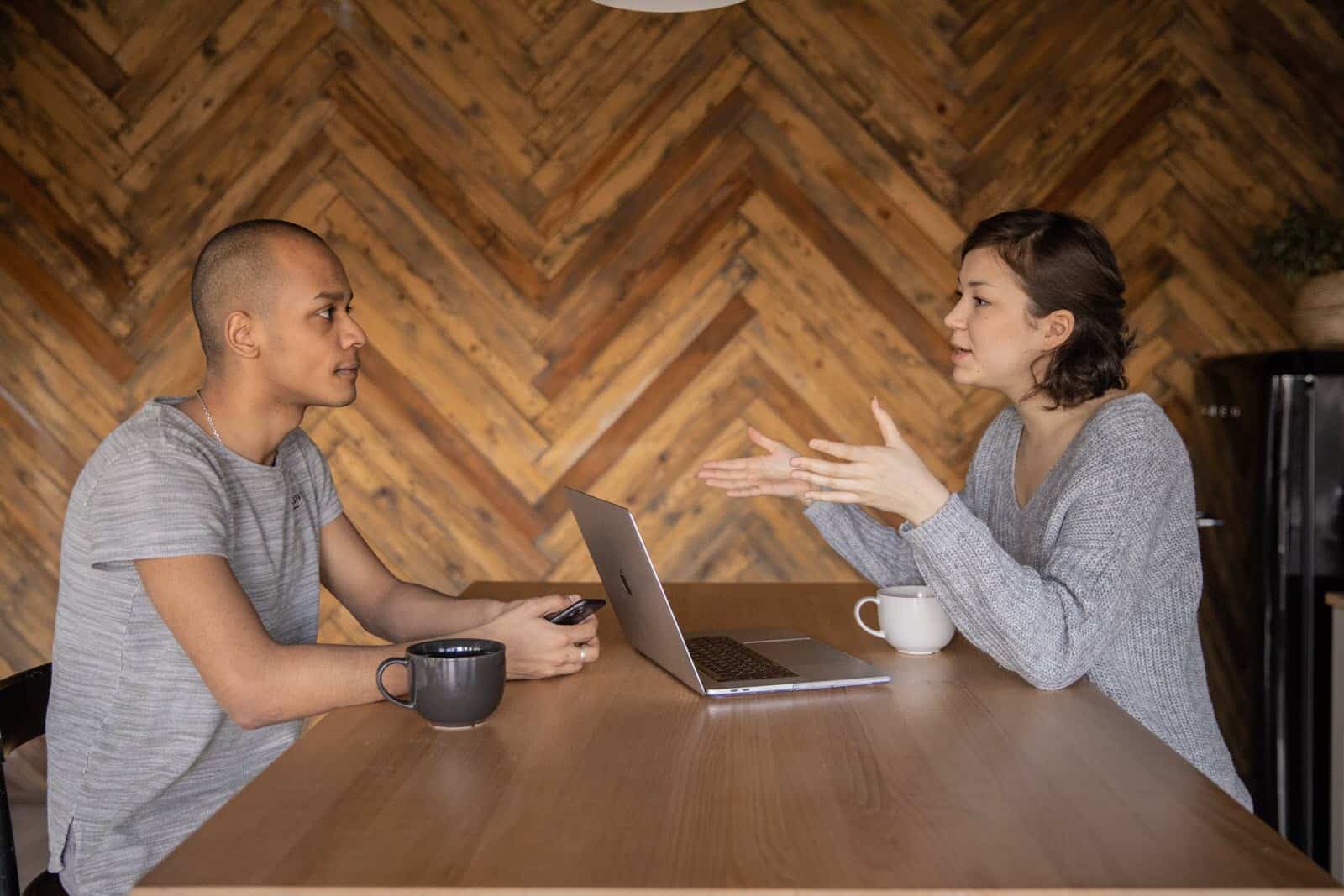 two people in discussion seated at a table, with coffee, phone, laptop